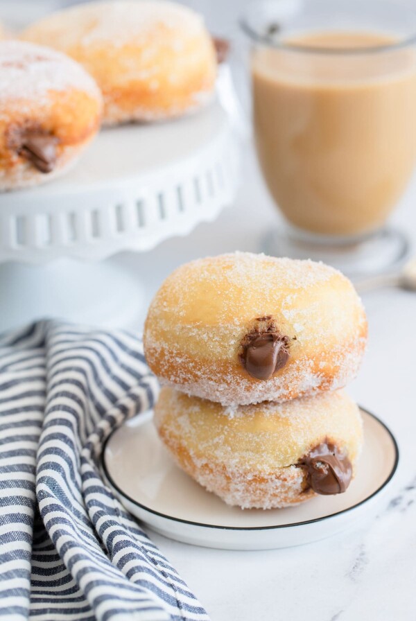 two nutella stuffed donuts on a small white plate with a blue and white towel on the side, a coffee and more donuts in the background.