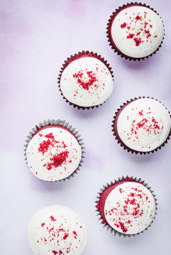 Six red velvet cupcakes with cream cheese frosting and cupcake crumbs on top.