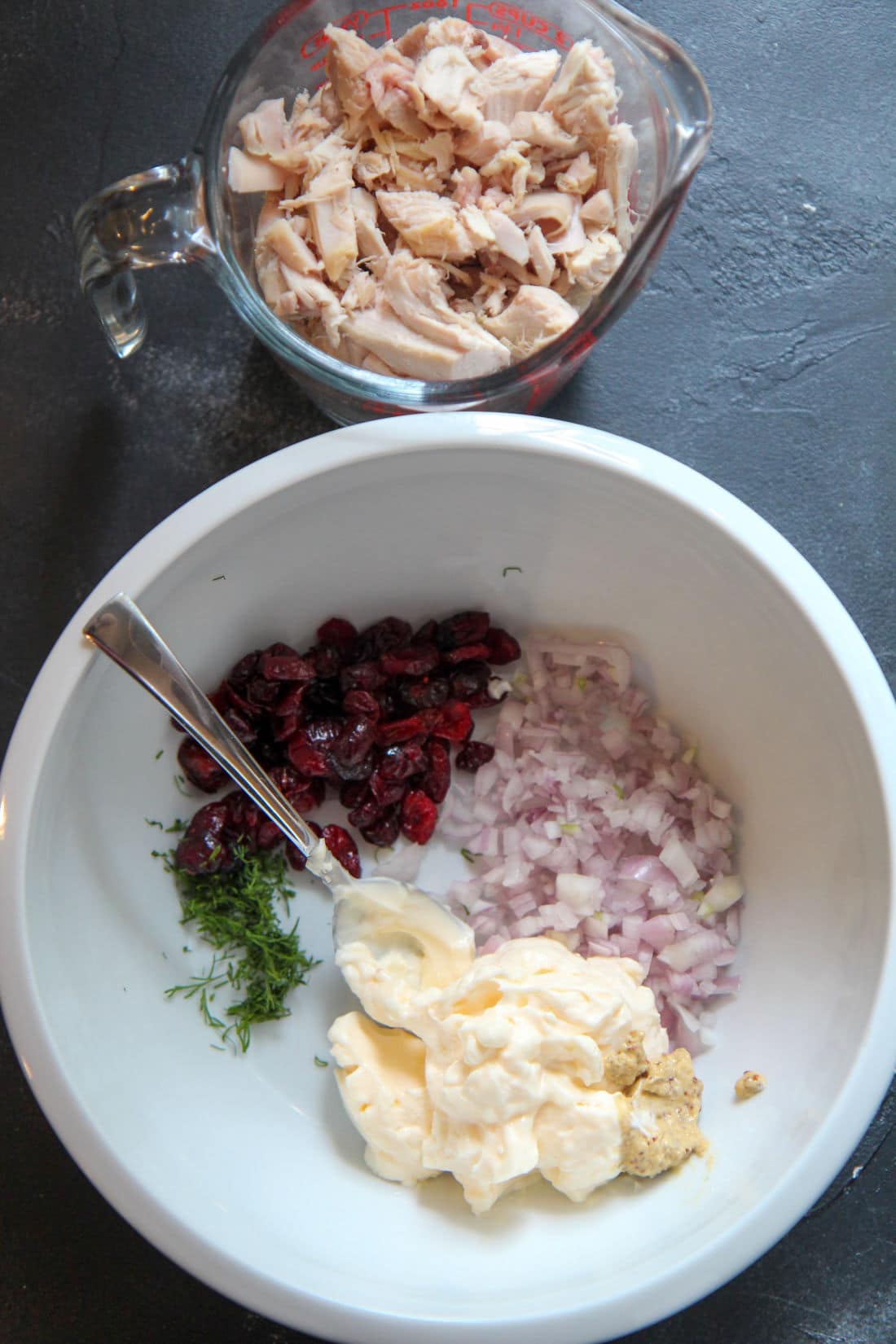 mayo, shallots, cranberries, and dill in a while bowl with a spoon and chicken in a cup