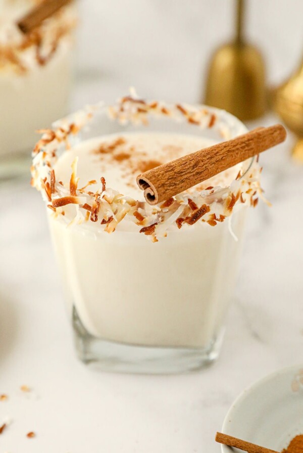 coquito in a short glass with toasted coconut on the rim and a cinnamon stick on top.