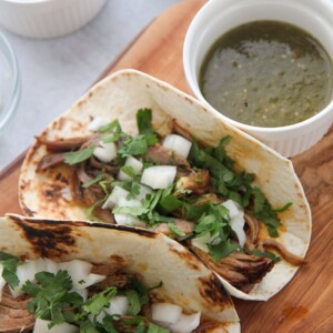 two instant pot pork carnitas tacos on a wooden board with green sauce on the side.