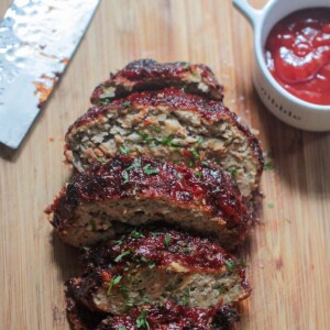 sliced meatloaf on a wooden board with a knife and ketchup on the side.