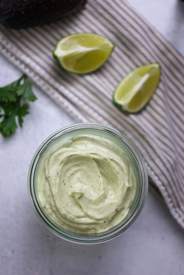avocado crema in a jar with limes, cilantro, and avocado in the background.