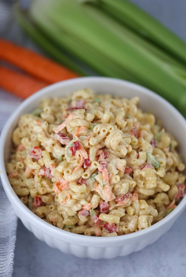 Deli-style macaroni salad in a white bowl with carrots and celery in the background.