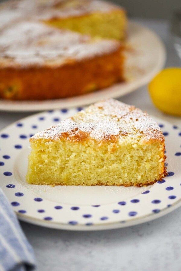 lemon olive oil cake slice on a plate with the entire cake in the background.