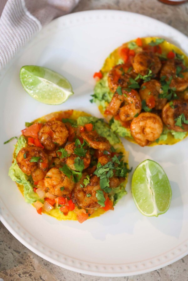 Two shrimp tostadas on a white plate with lime wedges on the side.