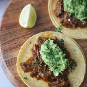 two shredded beef tacos on a wooden board with a lime wedge on the side.