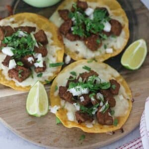 Three steak tacos with lime wedges and avocado crema on the side.