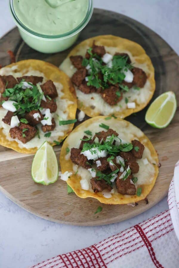 Three steak tacos with lime wedges and avocado crema on the side.