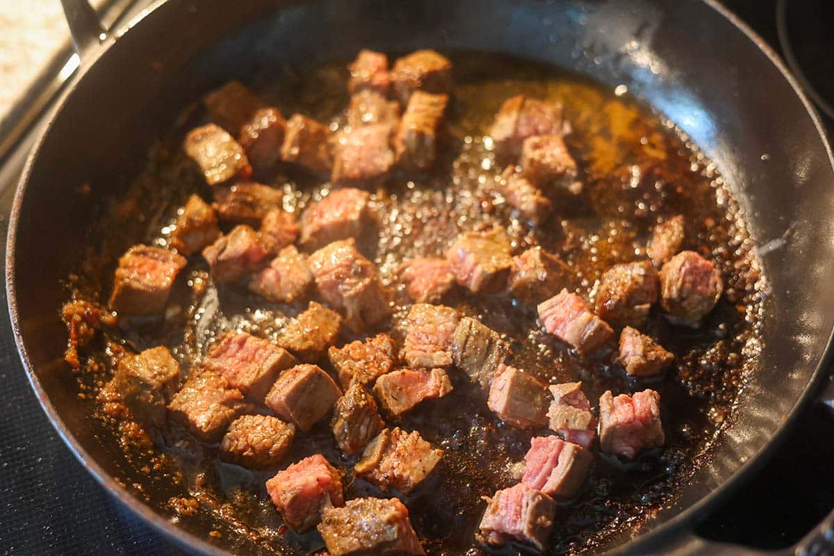 steak pieces cooking in a cast iron skillet on the stovetop. 